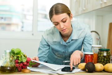 Young pensive housewife calculating family budget at kitchen..