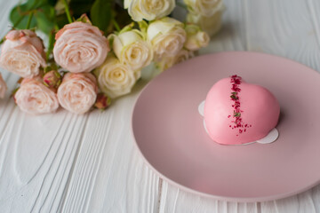 Heart-shaped pink cake with mirror glaze on pink plate with bouquet of small roses on a white wooden background. Congratulations on Valentine's Day, Happy Birthday, Women's Day. Elegant greeting card