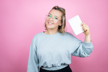 Young caucasian woman wearing sweatshirt over pink background smiling and showing blank notebook in her hand