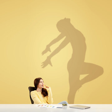 Young caucasian woman dreaming about future in big sport during her work in office. Shadow, silhouette of female dancer on the wall. Becoming a legend. Inspiration, aspiration. Copyspace.