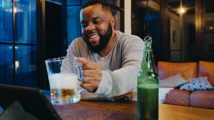 Middle-aged African American male drinking beer having fun happy night party event online celebration via video call in living room at home. Social distancing, quarantine for coronavirus prevention.