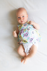 A baby girl lying on a bed on linen wearing a white cloth, reusable diaper and a pretty dress with...