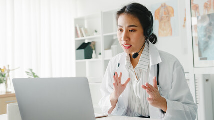 Young Asia lady doctor in white medical uniform with stethoscope using computer laptop talking video conference call with patient at desk in health clinic or hospital. Consulting and therapy concept.