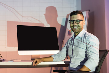 happy businessman in glasses looking at camera while sitting near computer monitor with blank screen