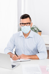 businessman in medical mask and glasses looking at camera while holding pen near notebook and laptop on desk