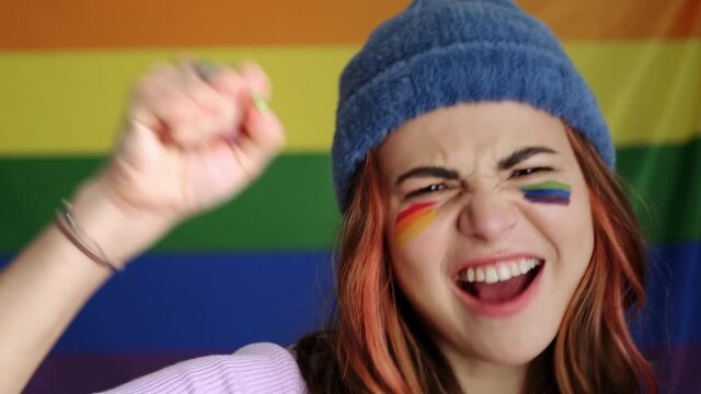 An emotional young woman is moving her hand to the side while opening her mouth and screaming standing isolated over rainbow flag