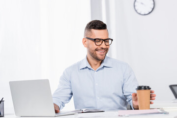 Obraz premium cheerful businessman in glasses reaching paper cup near laptop in office