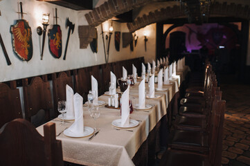 Table setting in the restaurant. White tablecloth, napkins, glasses, plates and knives with forks in a tavern with shields and swords of the knights. The concept of preparation for the celebration.