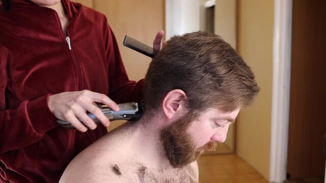 Adult bearded man gets haircut at home, during Coronavirus (Covid-19) isolation measures. Close up of Caucasian man's face while a woman uses a hair clipper to shorten his hair. 