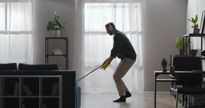 young man with headphones is washing floor in apartment, listening to music and dancing funny with mop