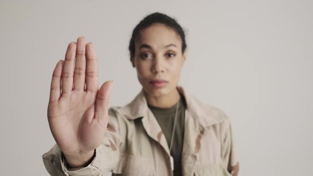 A serious young american soldier woman is showing stop gesture standing isolated over a gray background in the studio