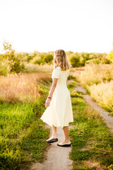 Fototapeta na wymiar A young beautiful girl with blond curly hair in a light summer yellow dress and with a straw hat in her hands in a field in the countryside at sunset. People and nature 