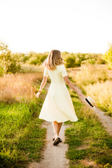 Fototapeta na wymiar A young beautiful girl with blond curly hair in a light summer yellow dress and with a straw hat in her hands in a field in the countryside at sunset. People and nature 