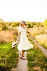 A young beautiful girl with blond curly hair in a light summer yellow dress and with a straw hat in her hands in a field in the countryside at sunset. People and nature 