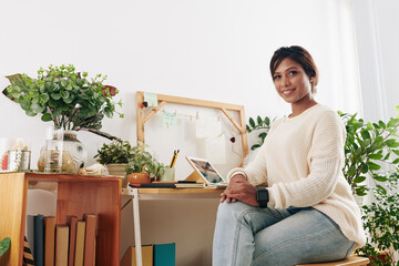 Fototapeta na wymiar Portrait of beautiful young Asian woman sitting at desk in her home office with plants around and looking at camera