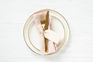 Golden rim plates and golden cutlery on pink napkin with tag on white table. Top view.
