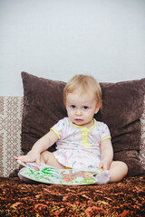 Fototapeta premium A little one-year-old girl is sitting on the sofa and looking at a picture book