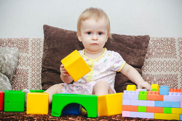A little one-year-old girl is sitting on the sofa and playing with cubes