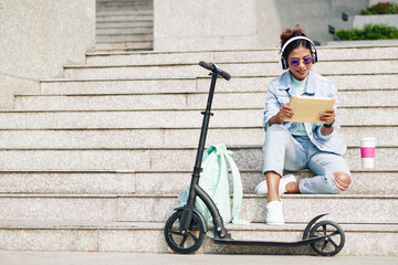 Happy young pretty woman sitting on steps after riding on electric scooter on campus and watching webinar on table computer © DragonImages