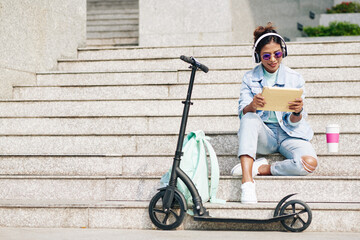 Happy young pretty woman sitting on steps after riding on electric scooter on campus and watching webinar on table computer