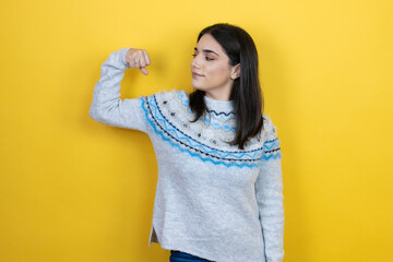 Young caucasian woman wearing casual sweater over yellow background showing arms muscles smiling proud