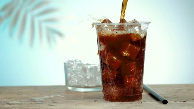 fresh cola drink poring in plastic take away cup with ice cubes forming foams and splashes, defocused palm leaf in background. slow motion