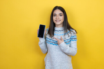 Young caucasian woman wearing casual sweater over yellow background holding smartphone showing screen smiling happy pointing with hand and finger