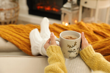 Woman with cup of hot drink resting on sofa near fireplace in living room, closeup