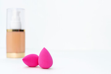 Close-up of clean pink sponge on the background of a bottle of beige cosmetic foundation for make up. Makeup artist accessories, products for perfect face skin on a white table. Copy space.
