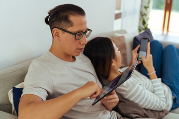 30s young adult Asian man and woman playing a mobile phone together sits on sofa in cozy living room at home. Happy couple internet user in casual clothes on couch. Technology usage in Asia concept