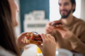 Unrecognizable young couple in love eating hamburgers indoors at home.