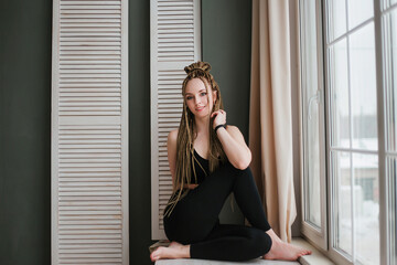 Fototapeta premium Young woman with long braids in sportswear practices yoga. A beautiful girl is sitting near a large window.
