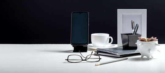 Home Office workspace. Desktop with smart phone template blank, notepads pens office suppliers cup of coffee drink plant. White work desk table on black background. Long web banner with copy space.