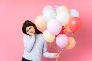 Young Ukrainian teenager girl holding lots of balloons over isolated pink background unhappy and frustrated