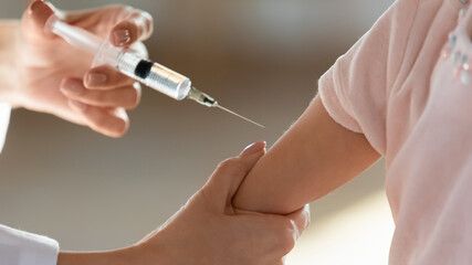 Crop close up of female nurse hold syringe make injection give vaccine to small child patient at checkup in hospital. Woman doctor vaccinate prick arm of little kid in clinic. Vaccination concept.