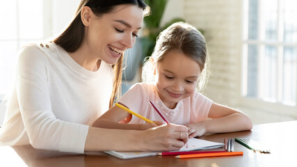 Overjoyed young Caucasian mother and little 7s daughter have fun drawing in album on family weekend together. Happy mom and small girl child engaged in funny activity paining. Hobby concept.