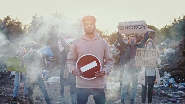 Portrait of Young Man Activist in Gas Mask Holding Stop Sign. In the Background People Protesting Against Garbage Pollution. Activists Standing in Toxic Smoke in a Garbage Dump. 