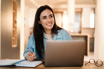 Happy charming woman writing down notes while working with laptop
