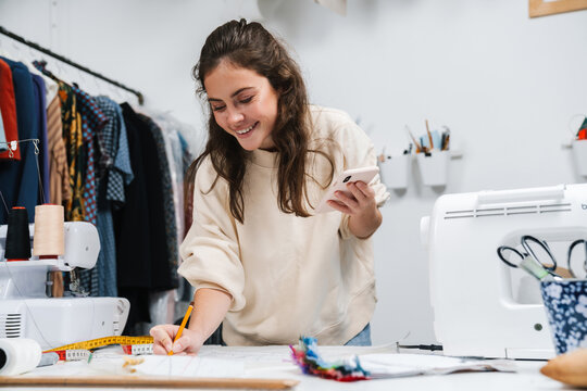 Smiling Girl Seamstress Using Mobile Phone While Working In Atelier