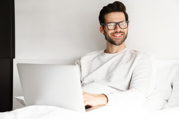 Smiling young man using laptop while resting in bed at home