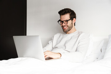 Smiling young man using laptop while resting in bed at home