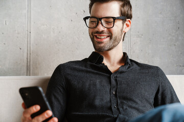 Smiling unshaven man using smartphone while sitting on couch