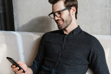 Smiling unshaven man using smartphone while sitting on couch