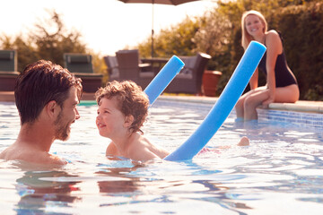 Family In Outdoor Pool On Summer Vacation Teaching Son To Swim With Noodle Swimming Aid