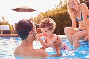 Family With Young Son Having Fun On Summer Vacation Splashing In Outdoor Swimming Pool