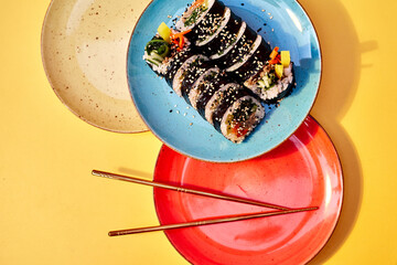 Delicious traditional Japanese food with sushi rolls. On a yellow background. Served on a blue plate with chopsticks. Sushi with rice, sesame, fish and vegetables