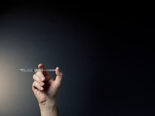 Fototapeta premium Colour image of man's hand holding syringe horizontally with liquid droplet on needle point, with dramatic lighting and text space to right
