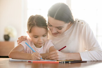 Loving happy young Caucasian mother and small daughter sit at table at home draw in album together. Smiling caring mom or nanny have fun play paint with little girl child. Hobby concept.