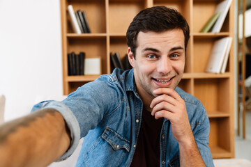 Joyful brunette young man smiling while taking selfie photo at home