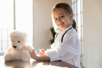 Portrait of smiling little 7s Caucasian kid act as doctor cure treat fluffy toy give injection or vaccine. Happy small girl child in white medical uniform play in hospital with teddy bear patient. - obrazy, fototapety, plakaty